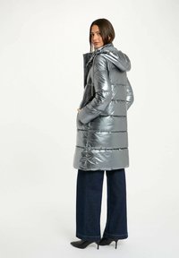 Grå metallic pufferjakke med hætte, der har vertikale quiltede sektioner og lynlås foran. Sammen med mørke brede jeans og høje hæle.