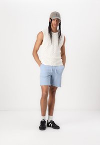 Pull sans manches blanc en tricot, short bleu clair, baskets noires avec accents blancs, casquette grise, pose debout sur fond blanc.