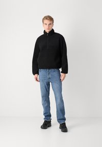 Zwarte fleece ritstrui met een hoge kraag, gedragen met lichtblauwe jeans en zwarte veterschoenen. Eenvoudige, casual outfit.