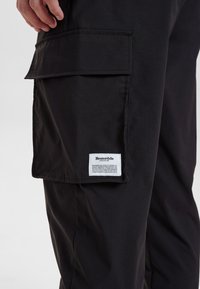 Zwarte cargobroek gemaakt van duurzaam materiaal; heeft een grote zijzak en een merketiket. Glad materiaal met zichtbare stiknaaddetails.