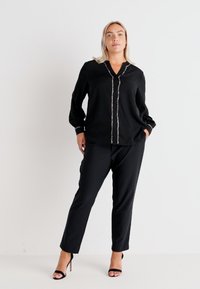 Blusa preta de manga longa com frente abotoada e detalhes brilhantes, combinada com calças pretas de perna reta e saltos altos pretos.