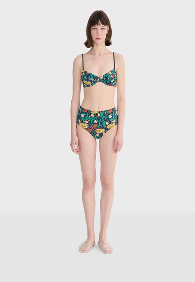 Femme debout portant un bikini taille haute vert à motifs floraux avec de fines bretelles et des chaussures transparentes à enfiler, sur un fond clair uni.
