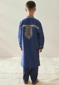 Camicia lunga blu navy con ricami d'oro intricati, caratterizzata da un colletto alto e maniche lunghe, abbinata a pantaloni coordinati e sandali marroni.