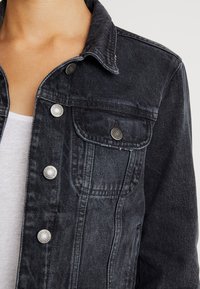 Zwarte denimjas met klassieke kraag, voorzak en zilveren knoopaccenten. Versleten textuur langs de naden. Draagt over een wit overhemd.