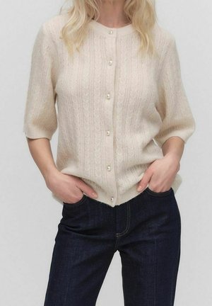 Cardigan tricoté couleur crème avec une texture côtelée, manches courtes et fermeture à boutons en perles, associé à un jean en denim foncé.