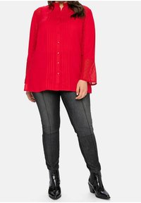 Blouse rouge à manches longues avec détails plissés et fermeture par boutons, associée à un jean gris foncé et des bottines noires, avec une finition élégante.