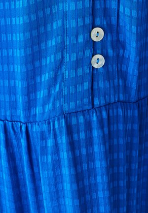 Modrá šatočka s vzorom a textúrovanou tkaninou. Má tierovanú konštrukciu a dva okrúhle krémové gombíky pozdĺž prednej strany.