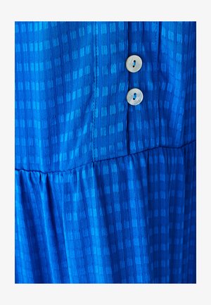 Modrá šatočka s vzorom a textúrovanou tkaninou. Má tierovanú konštrukciu a dva okrúhle krémové gombíky pozdĺž prednej strany.