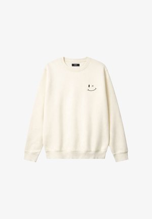 Sahnefarbener Pullover mit Rundhalsausschnitt und kleinem schwarzem Smiley-Gesicht, ein Auge offen und ein Auge durchgestrichen, auf der linken Brust.
