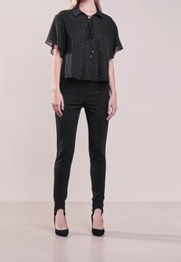 Blouse noire à manches courtes avec col et détail de cravate, assortie à un pantalon noir ajusté et des talons fermés, présentée sur un fond neutre.