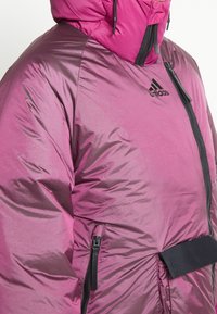 Lila puffjacka med blank yta, svarta dragkedjor, elastiska ärmslut och huva. Har en Adidas-logotyp på bröstet och sidoficka.