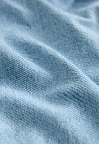 Gros plan d'un tissu denim doux bleu clair avec une texture de tissage visible et des plis légers créant des ombres et des reflets.