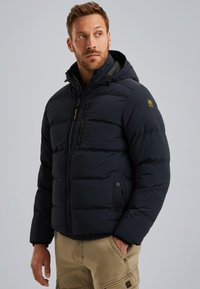 Padded navy jacket with a hood, zip front, side pockets, and a distinctive logo patch on the upper arm. Smooth texture and quilted design.