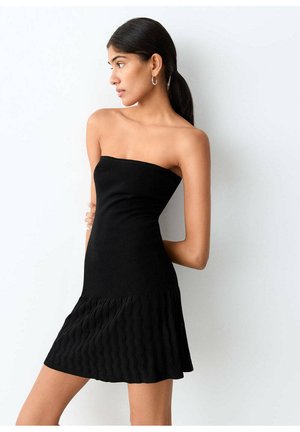 Femme aux cheveux foncés attachés en queue de cheval, portant des boucles d'oreilles créoles et une robe noire bustier avec un ourlet texturé, debout contre un mur blanc.