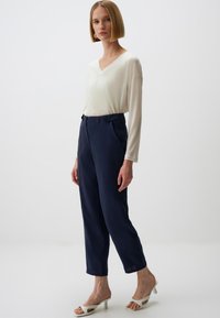 Weißes, langärmliges V-Ausschnitt-Oberteil, kombiniert mit marineblauen hochgeschnittenen Hosen. Das Model trägt weiße Absatzsandalen. Einfaches Design, glatte Textur.