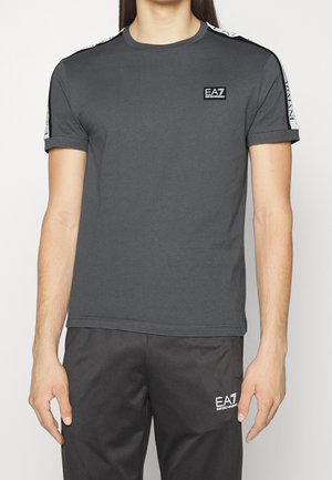 Homme portant un t-shirt EA7 Emporio Armani gris foncé et un pantalon noir avec des logos EA7 assortis sur la poitrine et la cuisse.