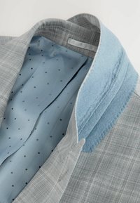 Grau gemusterter Blazer mit einem hellblauen Satinfutter, das kleine schwarze Punkte aufweist, und einem weichen blauen Filzkragen. Sichtbare Nahtdetails.