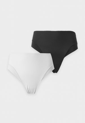 Femme aux longs cheveux foncés portant un soutien-gorge noir et un sous-vêtement taille haute marron foncé, debout devant un fond clair uni, regardant sur le côté.