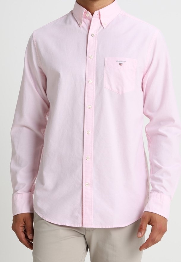 Uomo che indossa una camicia button-up a maniche lunghe di un rosa pallido con taschino e pantaloni beige, su uno sfondo semplice.