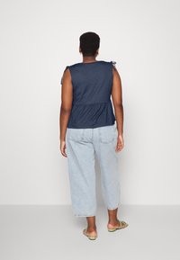 Top azul-marinho sem mangas com detalhes amarrados nos ombros, design peplum, combinado com jeans de perna larga azul-claro e sandálias verdes.
