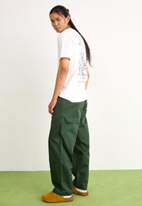 Tricou alb cu imprimeu grafic pe spate, asociat cu pantaloni verzi largi și saboți galbeni pe o suprafață verde deschis.