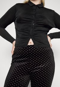 Top preto de mangas longas com um design justo e botões na frente, combinado com calças de veludo pretas de cintura alta com bolinhas prateadas.