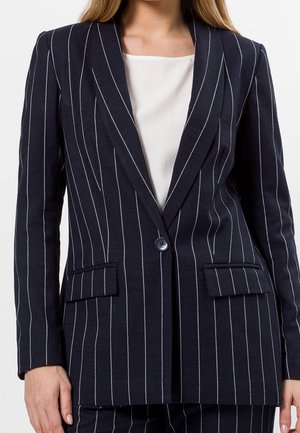 Vrouw draagt marineblauwe blazer met krijtstreep en enkele knoopsluiting, gecombineerd met bijpassende broek en een witte blouse eronder.