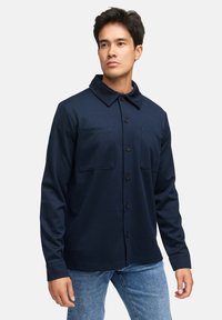 Mørkeblå skjorte med knapper lavet af glat stof, med krave og to brystlommer, parret med lyseblå denimjeans.
