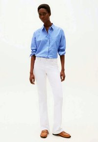 Woman wearing a blue striped button-up shirt, white high-waisted pants, and brown loafers, standing against a plain white background.