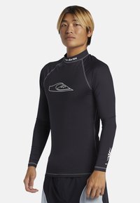 Černý dlouhý rukáv rash guard s přiléhavým střihem, šedými akcenty a malým bílým logem na hrudi. Vyrobeno z elastického materiálu.