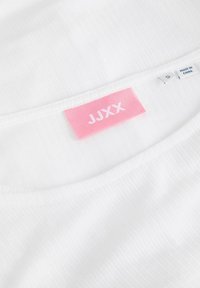 Encolure de vêtement blanc avec une étiquette de marque JJXX rose et une petite étiquette de taille indiquant "S" et "Fabriqué en Chine".