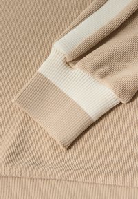 Tejido de suéter ribeteado en color beige claro con una franja de marfil contrastante en el puño. La vista en primer plano muestra la textura y el detalle del tejido.