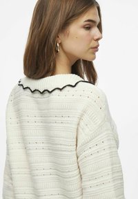 Svetr pletený v krémové barvě s texturovaným vzorem a černým krajkovým límcem. Nabízí volný střih a spadlé ramena, zdůrazňující složité detaily.