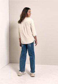 Camicia beige chiaro con maniche a tre quarti, dettaglio raccolto sul retro, abbinata a jeans in denim blu e scarpe da ginnastica bianche. Sfondo neutro.