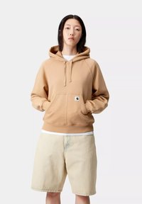 Sudadera marrón clara con un bolsillo tipo canguro, capucha con cordón y puños acanalados. Combinada con pantalones cortos beige. Textura suave, diseño casual.