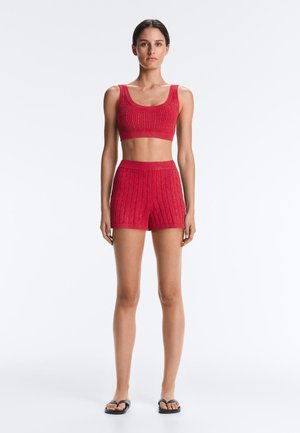 Vrouw staand met een rood gebreid crop top en bijpassende shorts met hoge taille, gecombineerd met zwarte teenslippers, op een effen witte achtergrond.