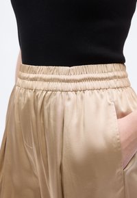 Personne portant un pantalon en satin beige avec une taille élastique et une poche latérale, associé à un haut côtelé noir, main dans la poche.