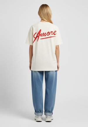 T-shirt oversize crème avec un script rouge "Amore" dans le dos, associé à un jean bleu clair, des baskets blanches et une texture lisse.