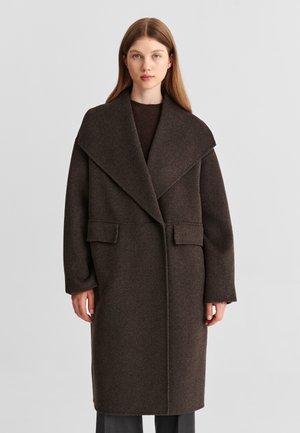 Jeune femme aux longs cheveux raides portant un manteau en laine oversize marron foncé avec de larges revers et des poches à rabat, debout devant un fond uni.