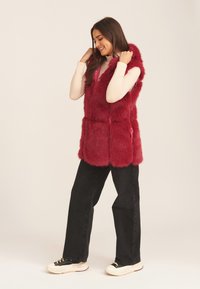 Red faux fur vest with a hood, front pockets, and a relaxed fit, paired with black pants and white sneakers. Soft texture and bold color.