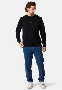 Svart sweatshirt med rund halsringning och vittryckt text. Ett par blå jeans och vita sneakers. Avslappnad passform med långa ärmar och ribbade muddar.