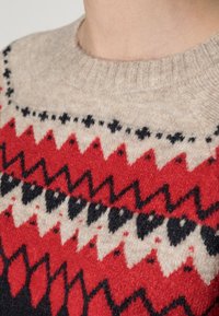 Villapaita, jossa on vaalea beige pohjaväri ja geometrinen kuviointi punaisena ja mustana, pyöreä pääntie sekä resorikuvio.