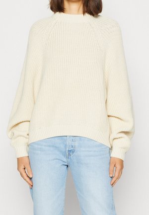 Žena v krémovém pleteném svetru s balónovými rukávy a světlých světle modrých džínách s vysokým pasem, stojící před jednobarevným pozadím.