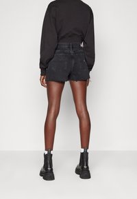 Person klädd i en svart kort sweatshirt, högmidjade svarta denimshorts med fransade kanter, vita strumpor och svarta klumpiga stövlar, står med ryggen vänd.