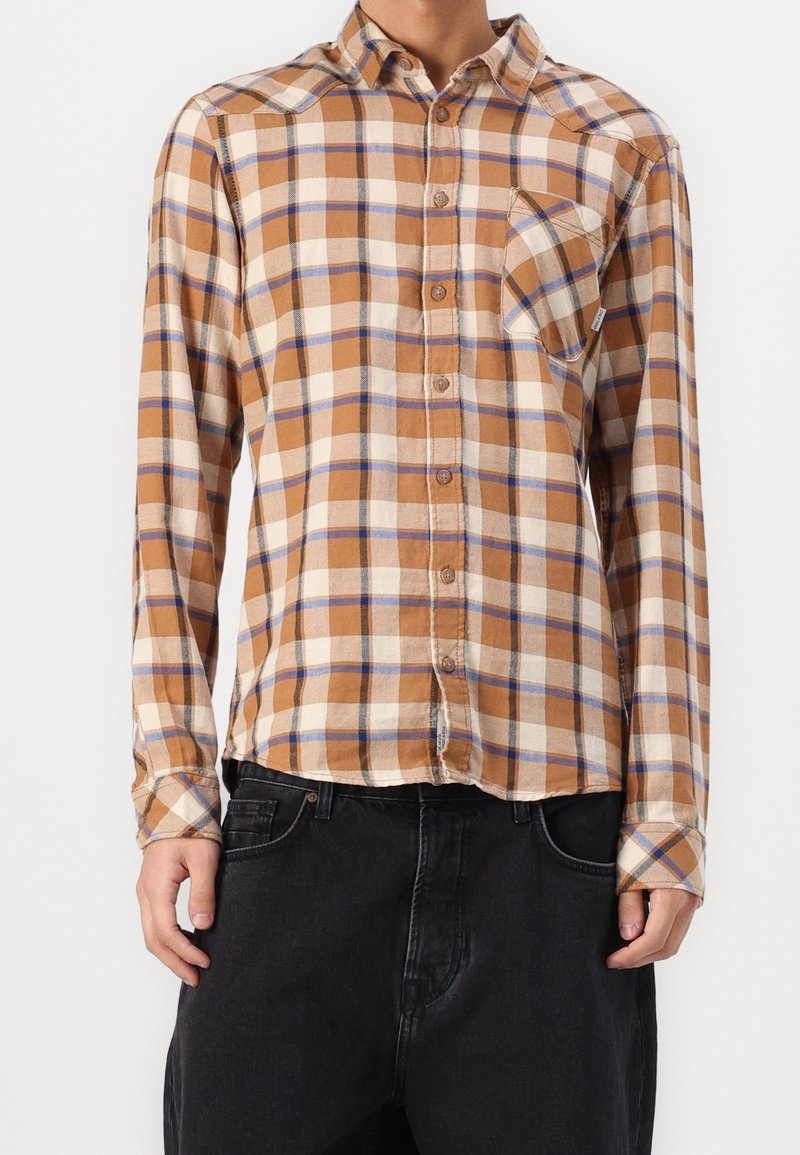 Personne portant une chemise à carreaux marron, beige et bleue à manches longues avec une poche avant et un pantalon noir, debout devant un fond uni.