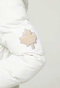 Witte pufferjack met een beige, gestructureerde esdoornbladpatch op de elleboog, met een gladde, glanzende buitenkant en een verzameld mouwontwerp.