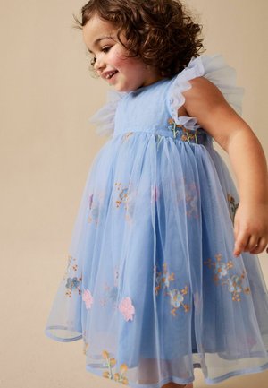 Lille pige smilende, iført en lyseblå kjole med flæseærmer og broderede blomsterdesigns, stående mod en neutral baggrund.