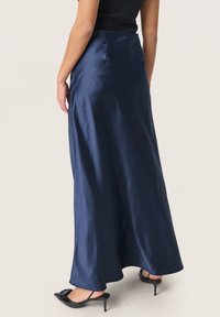 Lunga gonna in satin blu navy con una texture liscia e un design fluido, caratterizzata da una vita alta e uno spacco laterale, abbinata a scarpe nere con tacco.