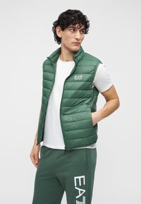 Muž oblečený v zelenej bezrukávovej prešívanej veste, bielom tričku a zelených teplákoch s logom EA7, stojaci s rukou v vrecku a pozerajúci sa bokom.