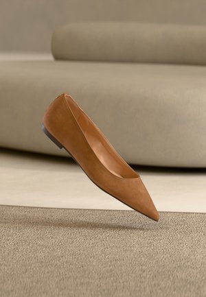 Brązowy płaski but z ostrym noskiem unoszący się nad teksturowanym beżowym dywanem, w tle jasnobeżowa, zakrzywiona sofa.